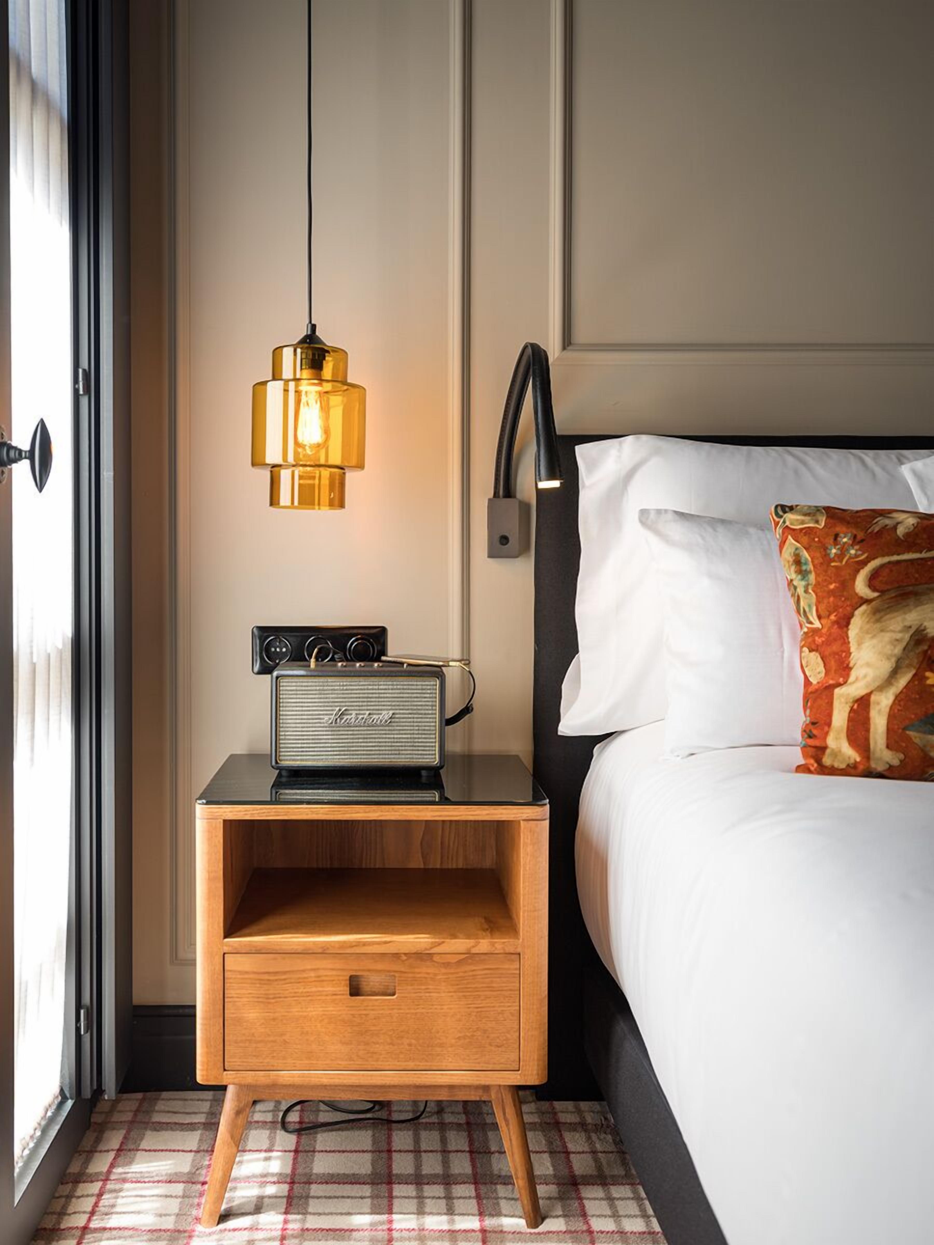 A neatly made bed with a white pillow and a decorative pillow with a dog design. A vintage radio is placed on a wooden nightstand beside the bed. A modern pendant light hangs above the radio.
