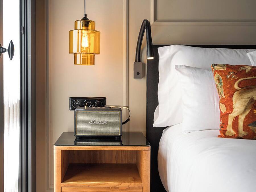 A neatly made bed with a white pillow and a decorative pillow with a dog design. A vintage radio is placed on a wooden nightstand beside the bed. A modern pendant light hangs above the radio.