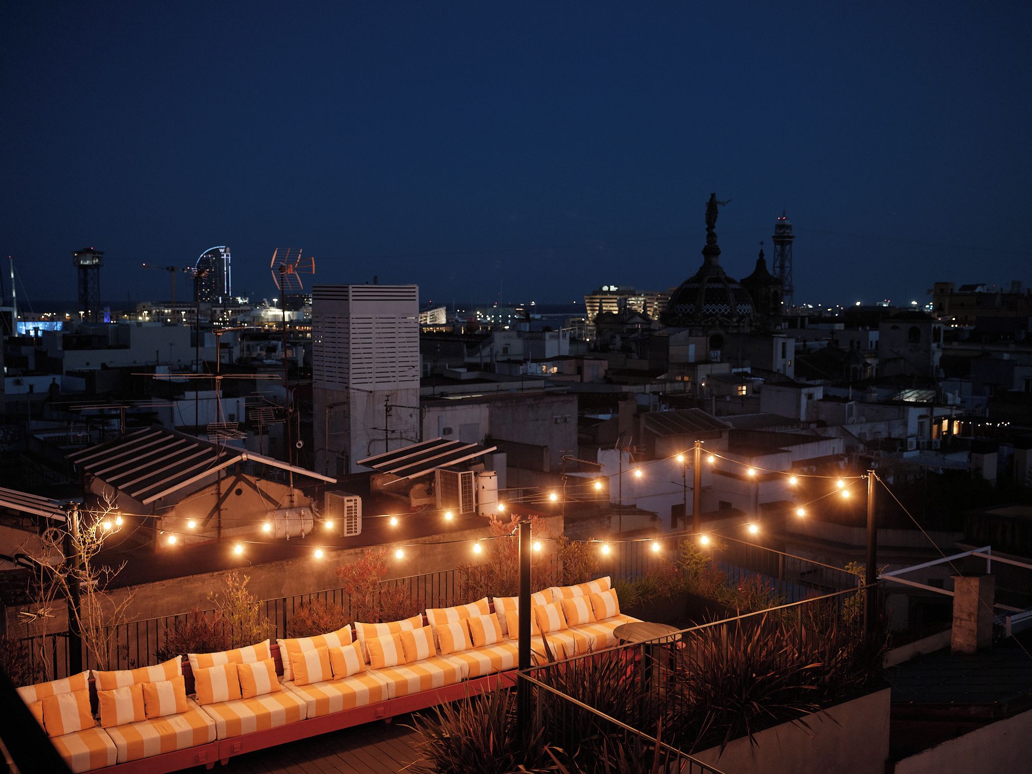 A rooftop terrace with a sofa and string lights, overlooking a cityscape at night.