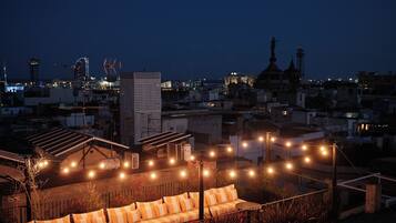 A rooftop terrace with a sofa and string lights, overlooking a cityscape at night.