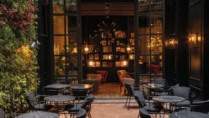 Un café en plein air avec des tables et des chaises, un mur végétal orné de plantes et une entrée d’immeuble avec des portes vitrées.