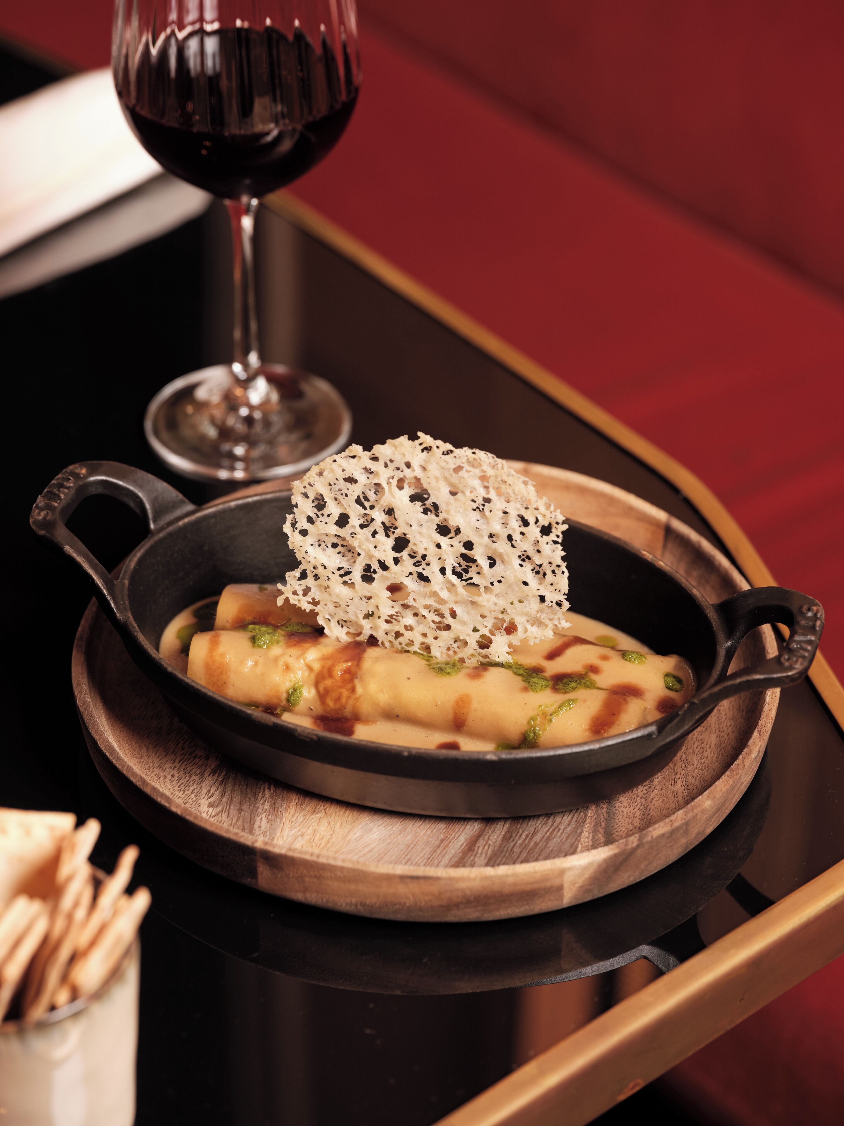 Un plato con preparaciones enrolladas en una sartén de hierro fundido, una copa de vino tinto y una mesa de madera.