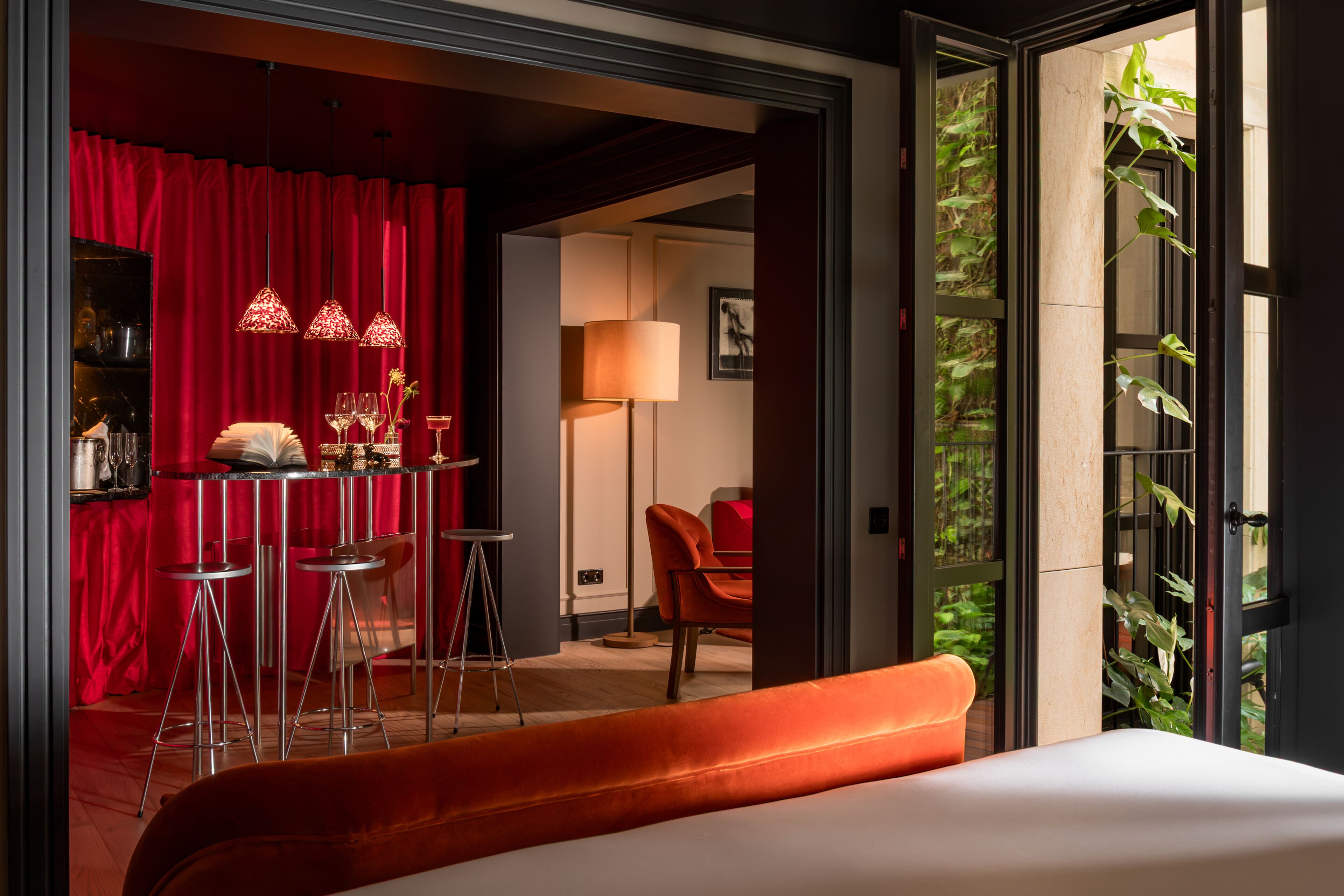 A hotel room with a red curtain, a bar area with stools, a table with glasses, and a bed with a white headboard.