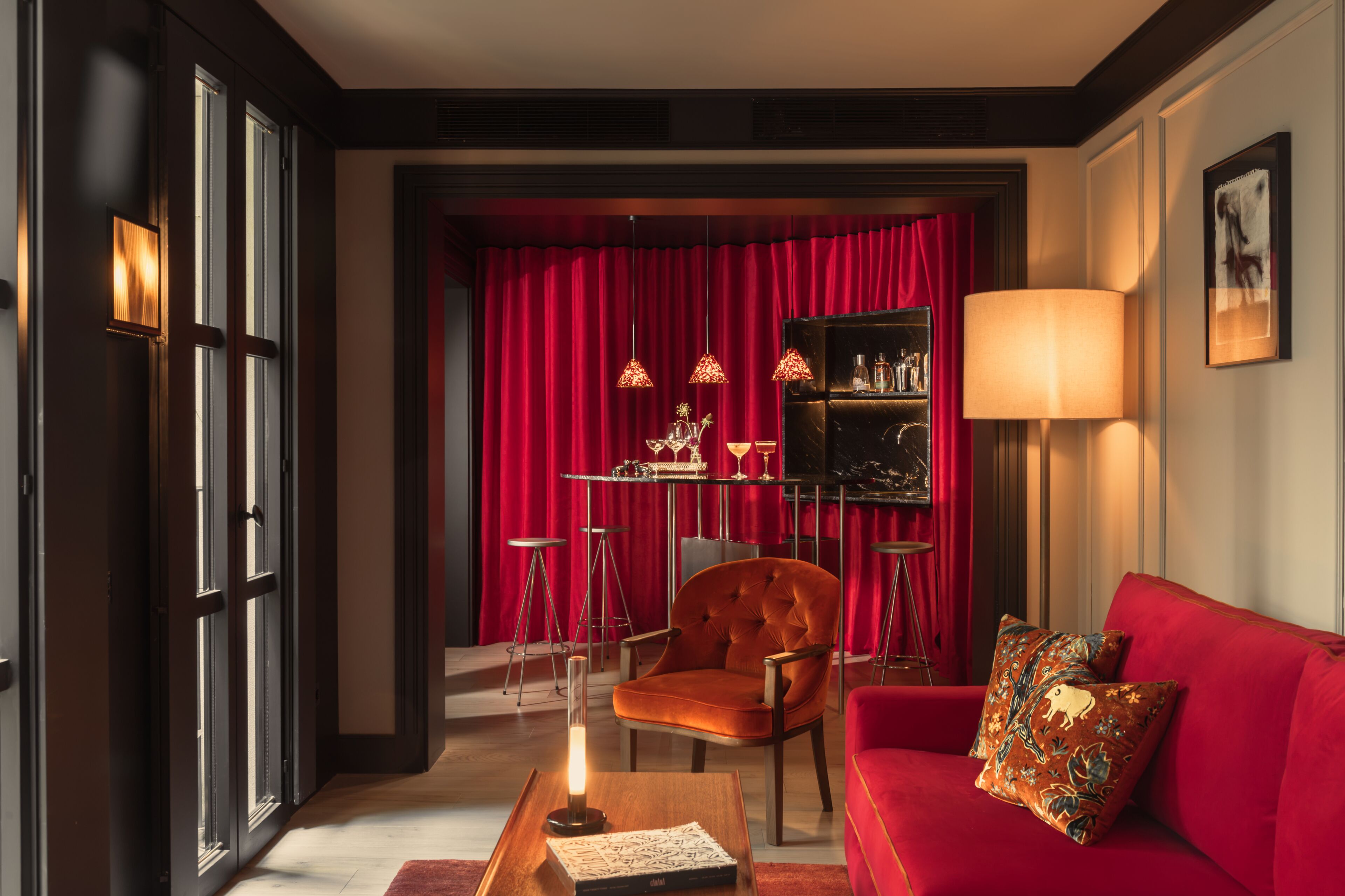 Una habitación con un sofá rojo, un telón rojo de fondo, una mesa con una lámpara y una silla.