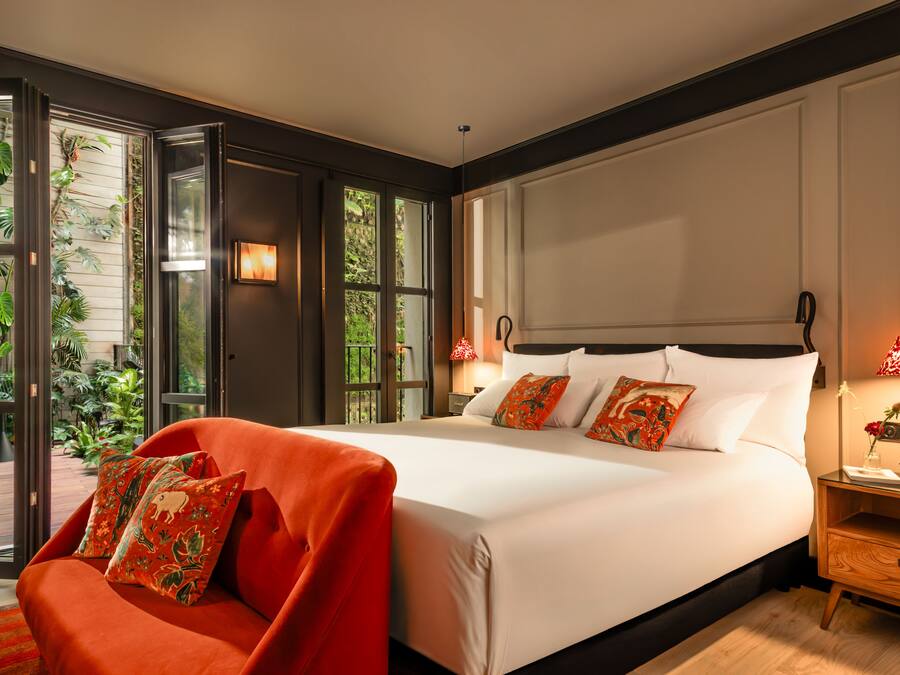 A modern hotel room with a large bed, a red armchair, and a view of greenery through the windows.