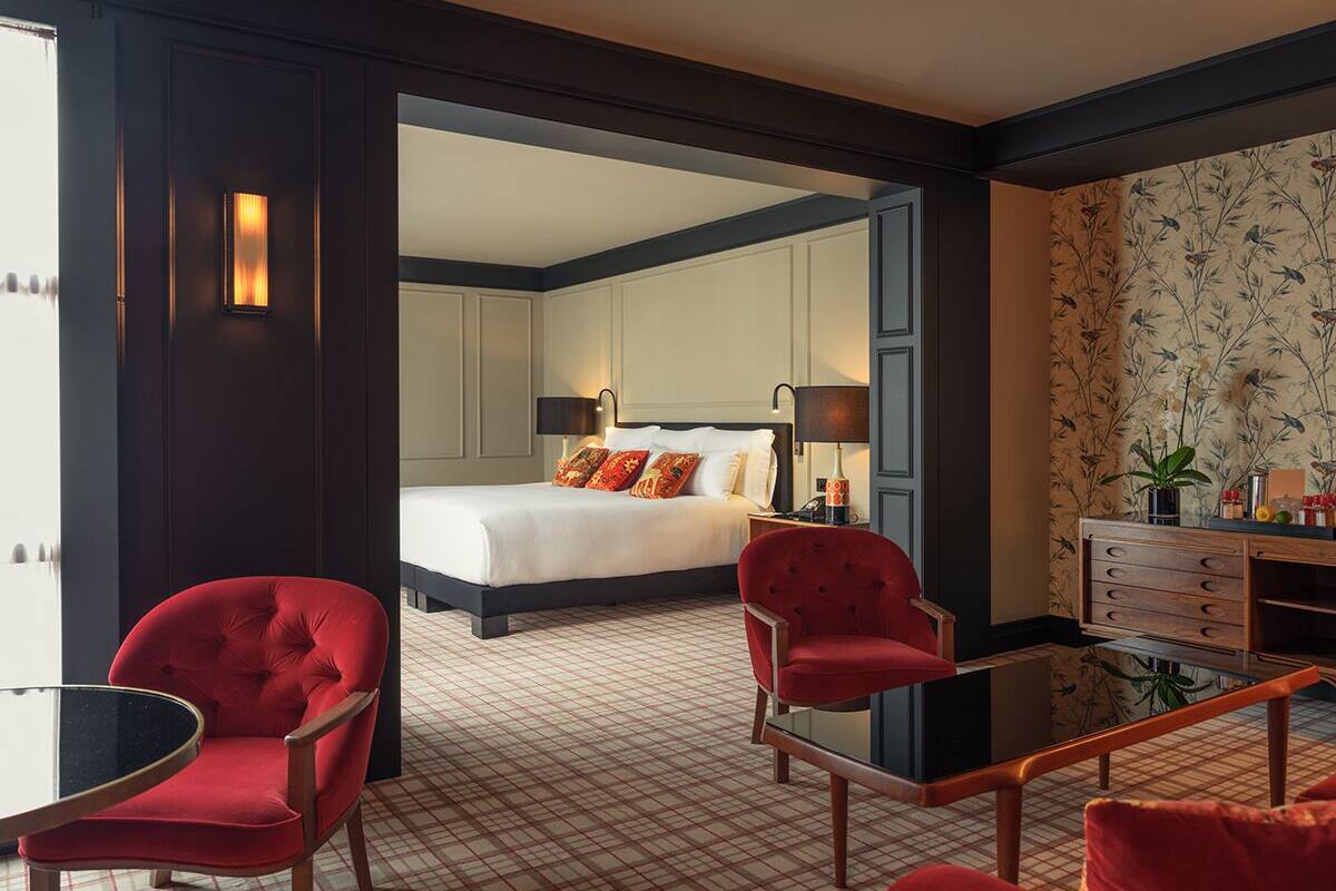 A hotel room with a large bed, two red armchairs, a small table, and a dresser with a mirror.