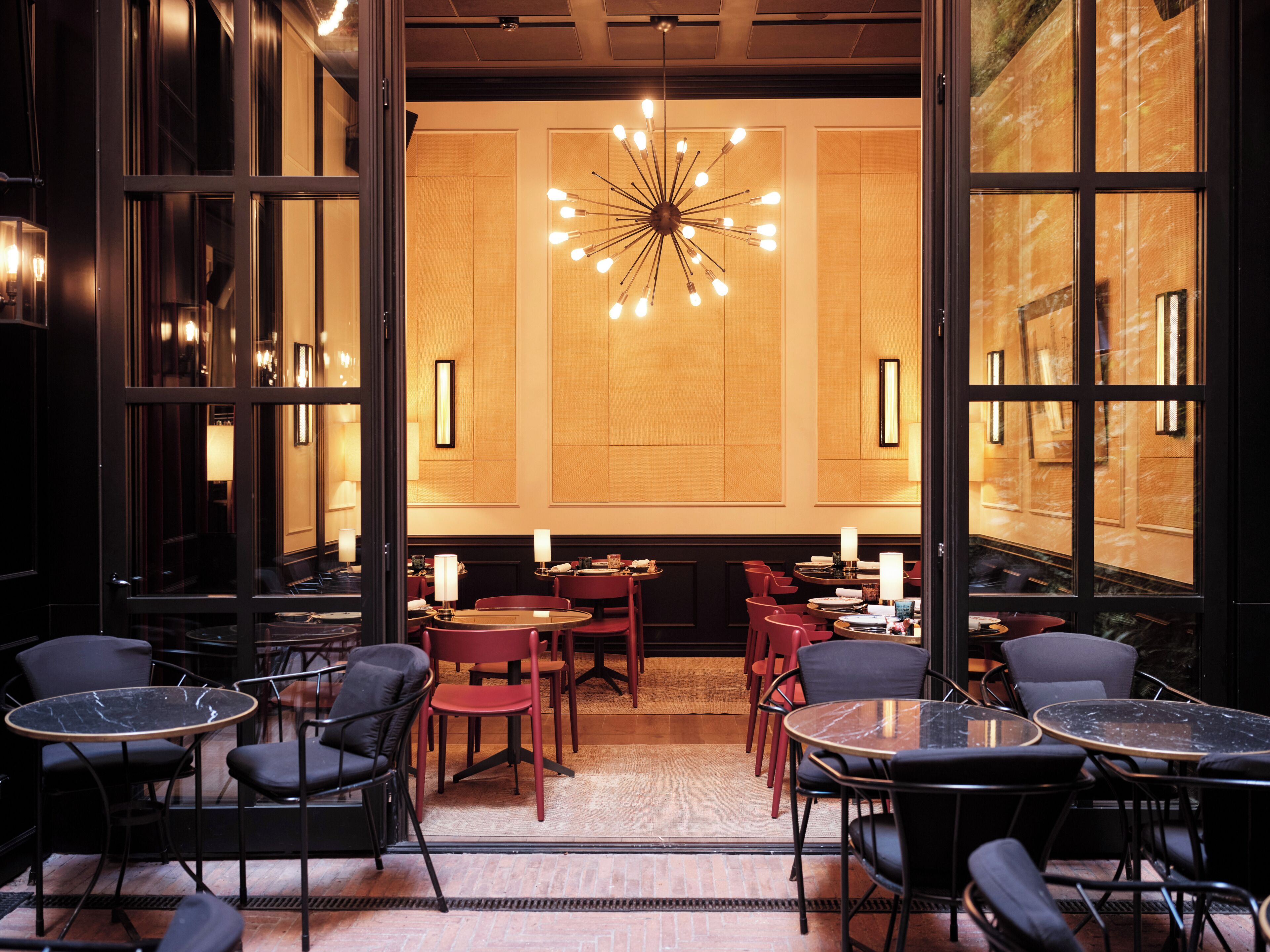 A modern dining area with round tables and chairs, a central chandelier, and large windows.