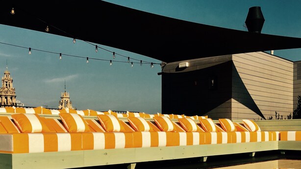 Une terrasse sur le toit, ornée d’une rangée de coussins rayés orange et blancs, d’une guirlande lumineuse, et avec un bâtiment moderne en arrière-plan.