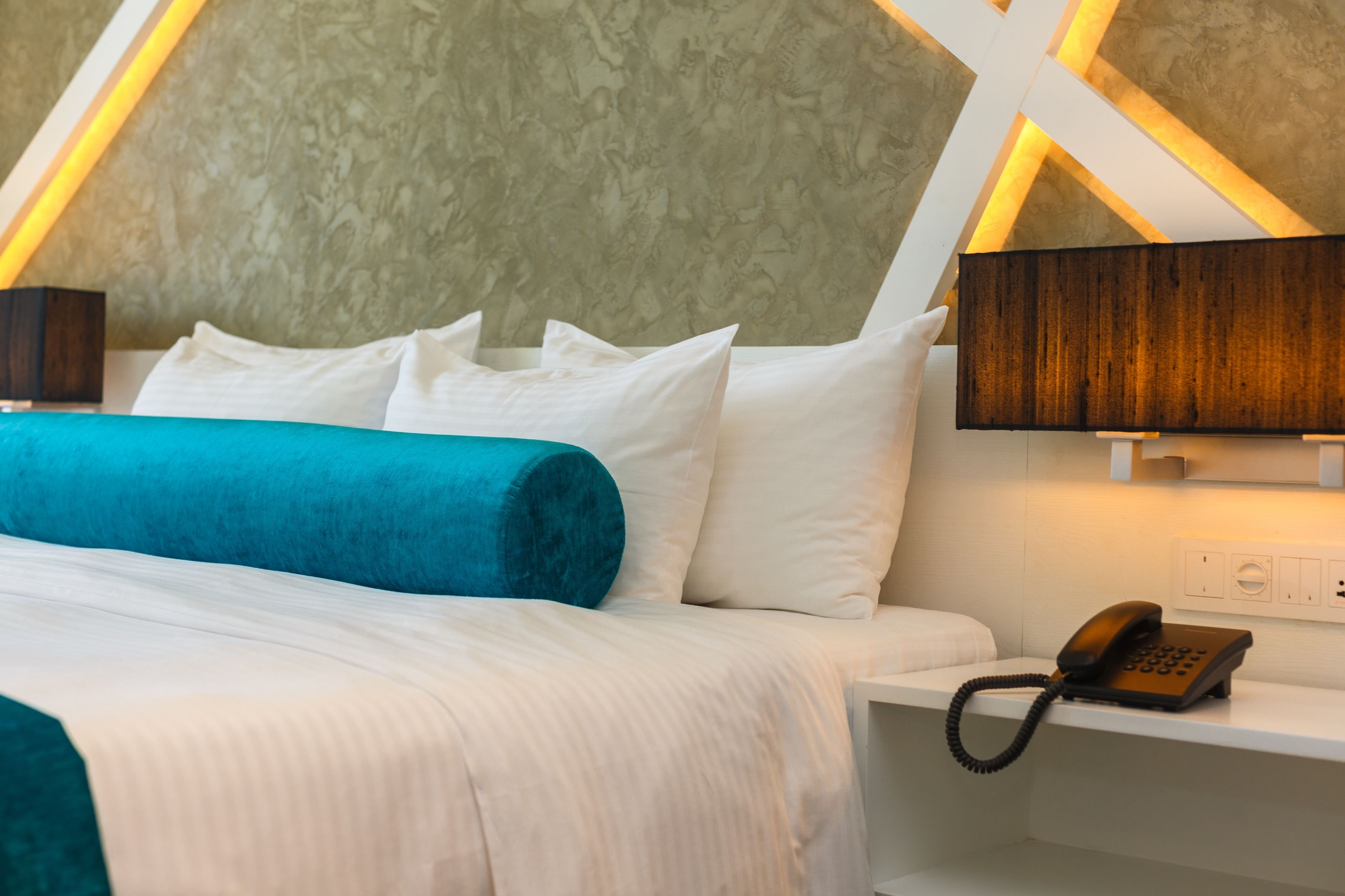 A hotel room with a bed, white bedding, a teal throw blanket, a bedside telephone, and a wall-mounted lamp.