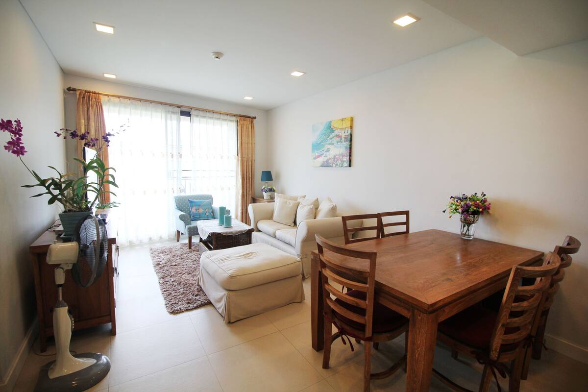 A living room with a wooden dining table, white sofa, and a small side table with a lamp.