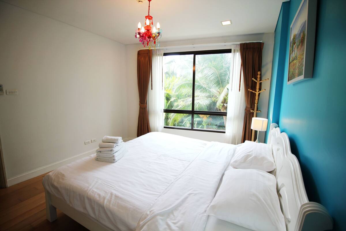 A neatly made bed with white linens, a chandelier, a window with curtains, and a wall-mounted lamp.