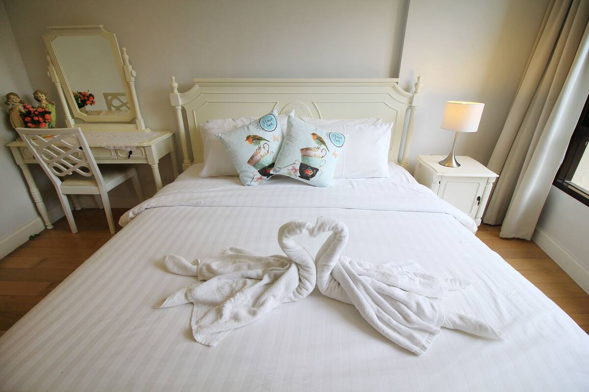 A neatly made bed with white linens, two decorative pillows, a bedside table with a lamp, and a chair with a mirror.
