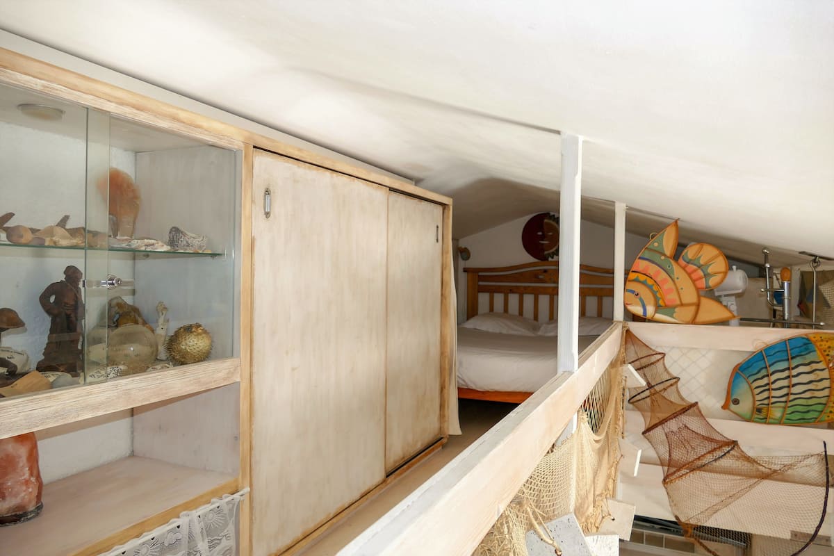 A room with a wooden wardrobe, a glass display cabinet with decorative items, a bed with a white mattress, and a wall with colorful fish decorations.