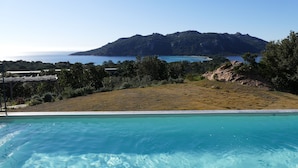 Una piscina con agua azul cristalina, rodeada de césped y árboles, con una montaña y el mar al fondo.