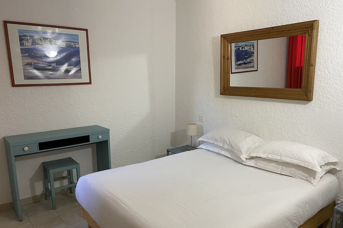 A bedroom with a bed, a desk, a chair, a nightstand, a lamp, and two framed pictures on the wall.