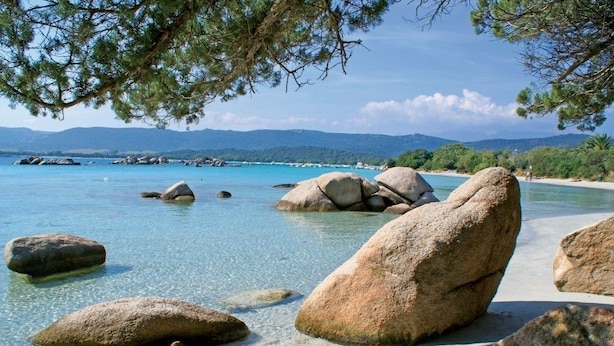 Una playa con rocas grandes y agua cristalina, rodeada de vegetación y montañas al fondo.