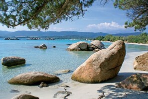 Une plage bordée de gros rochers et aux eaux cristallines, entourée de verdure et avec des montagnes en toile de fond.