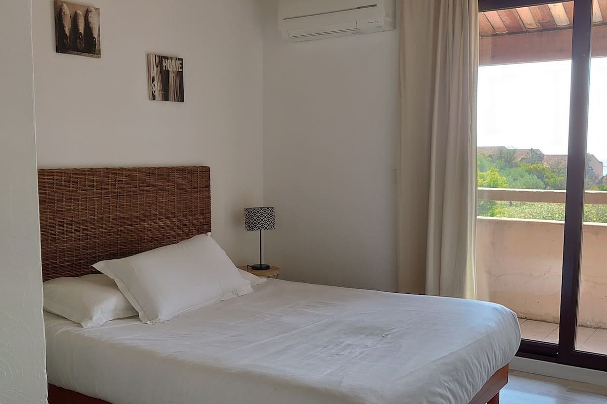 A bedroom with a wooden bed, a nightstand, an air conditioning unit, and a view through a large window.
