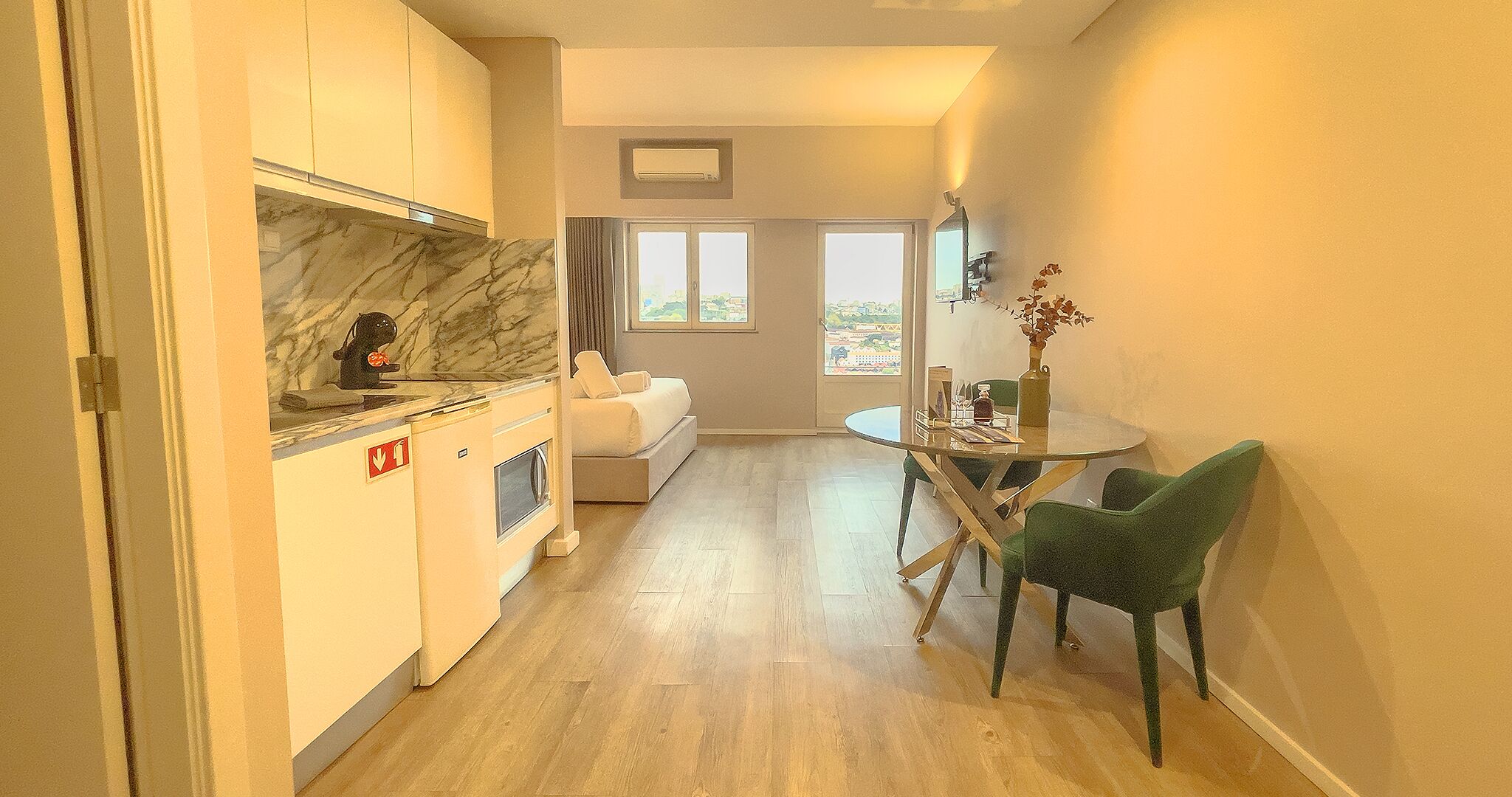 Una cocina compacta con encimera de mármol, un comedor con mesa de vidrio y sillas verdes, y vistas a través de una ventana.