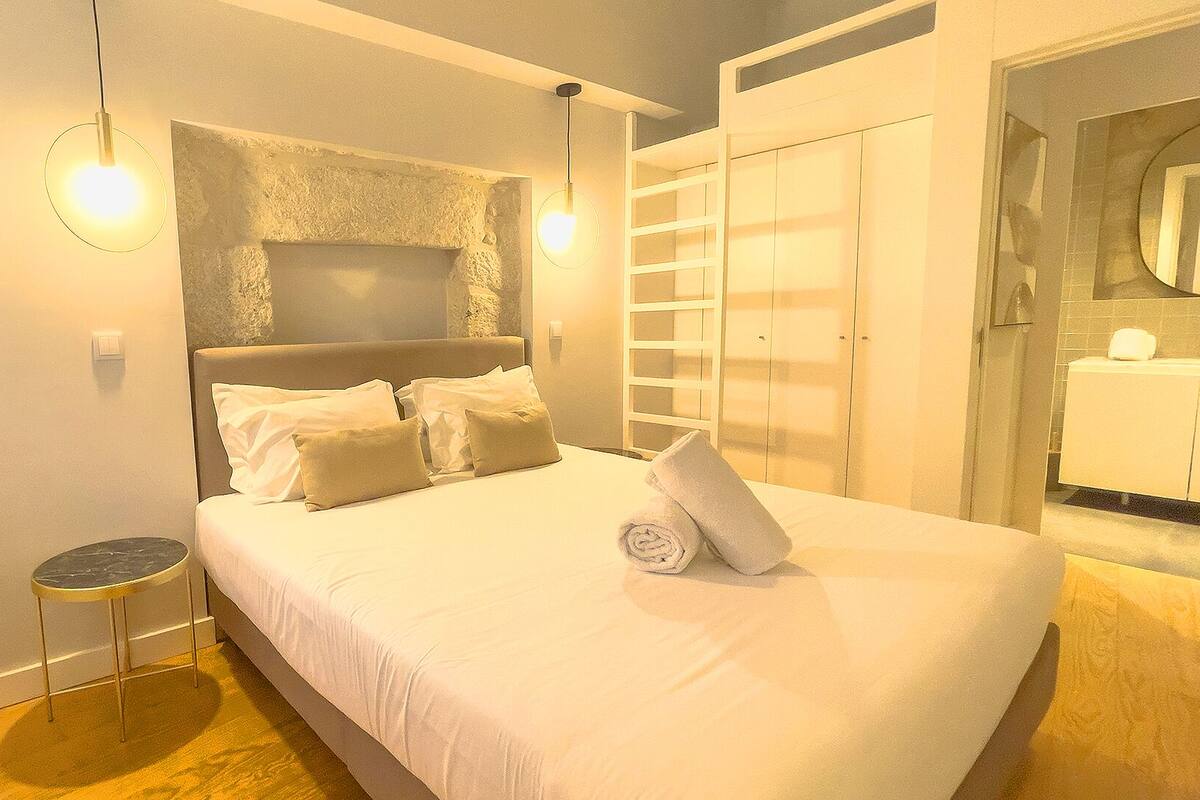 A bedroom with a bed, pillows, a round side table, a wall-mounted light fixture, and a wardrobe.