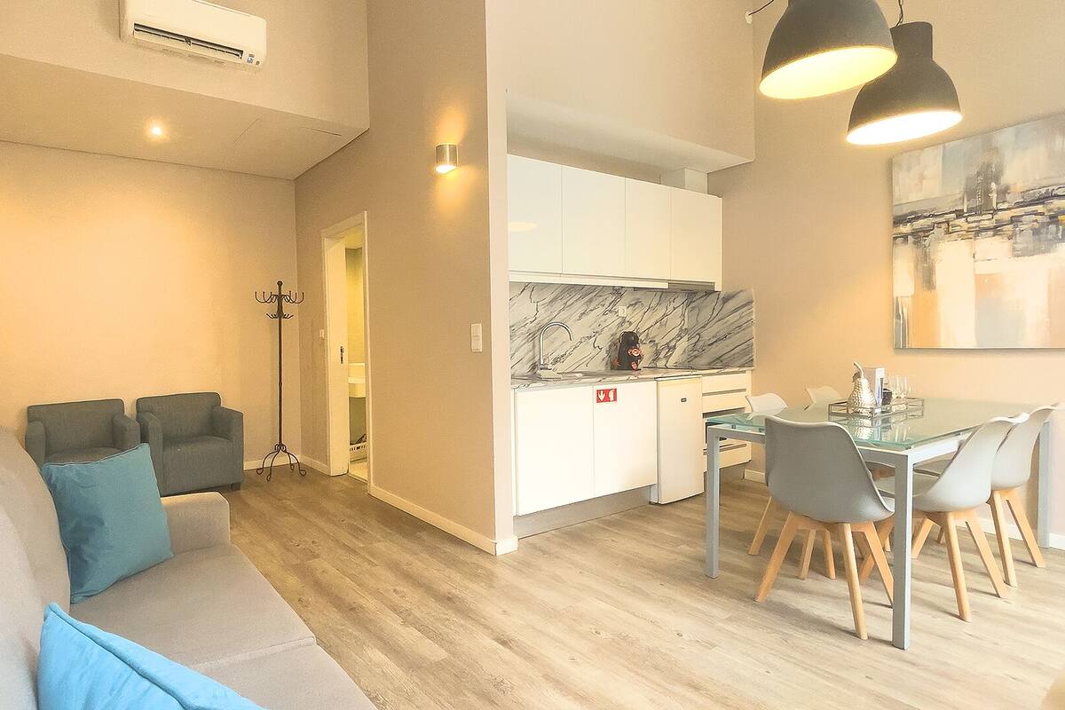 A modern living room with a sofa, a dining area with a table and chairs, and a kitchen with white cabinets and a marble backsplash.