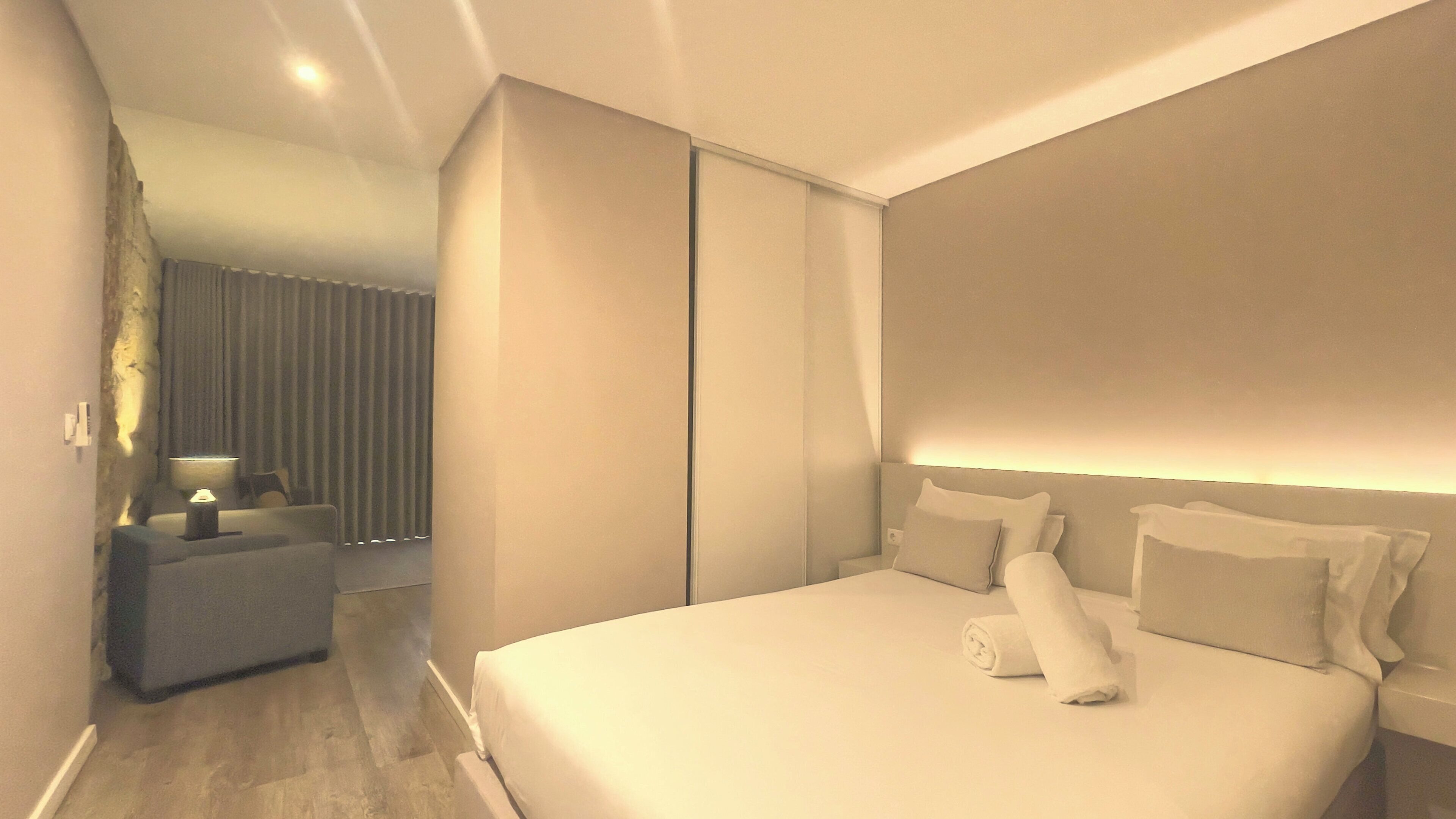 Una habitación de hotel moderna con una cama grande, una mesita de noche, una lámpara y vistas al pasillo.