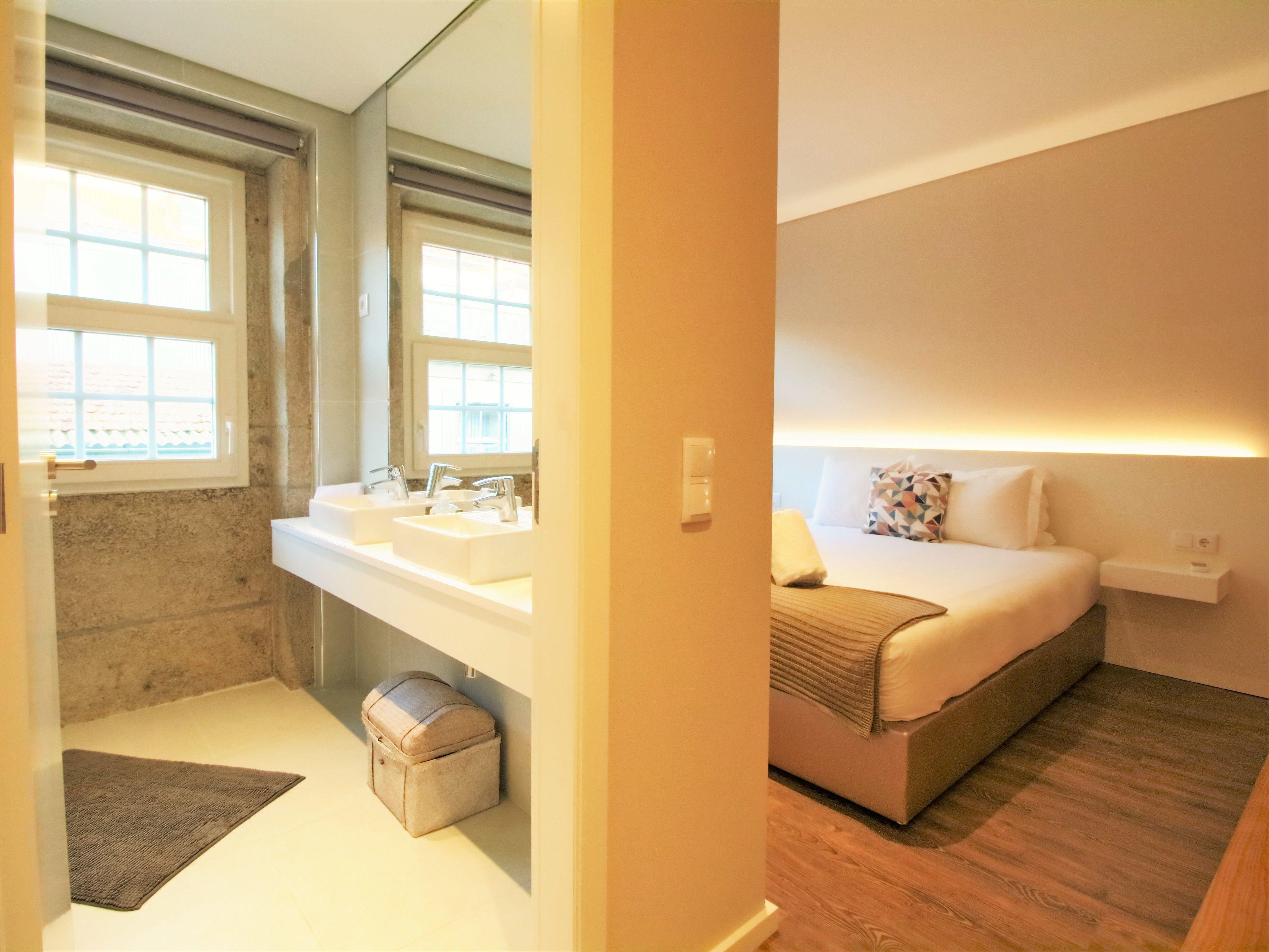 Ein Hotelzimmer mit Badezimmer, das zwei Waschbecken, einen Spiegel und ein Fenster zeigt. Das Zimmer verfügt über ein Bett mit Kopfteil, einen Nachttisch und eine Nachttischlampe.