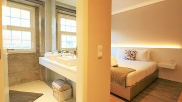 A hotel room with a bathroom featuring two sinks, a mirror, and a window. The room has a bed with a headboard, a bedside table, and a bedside lamp.