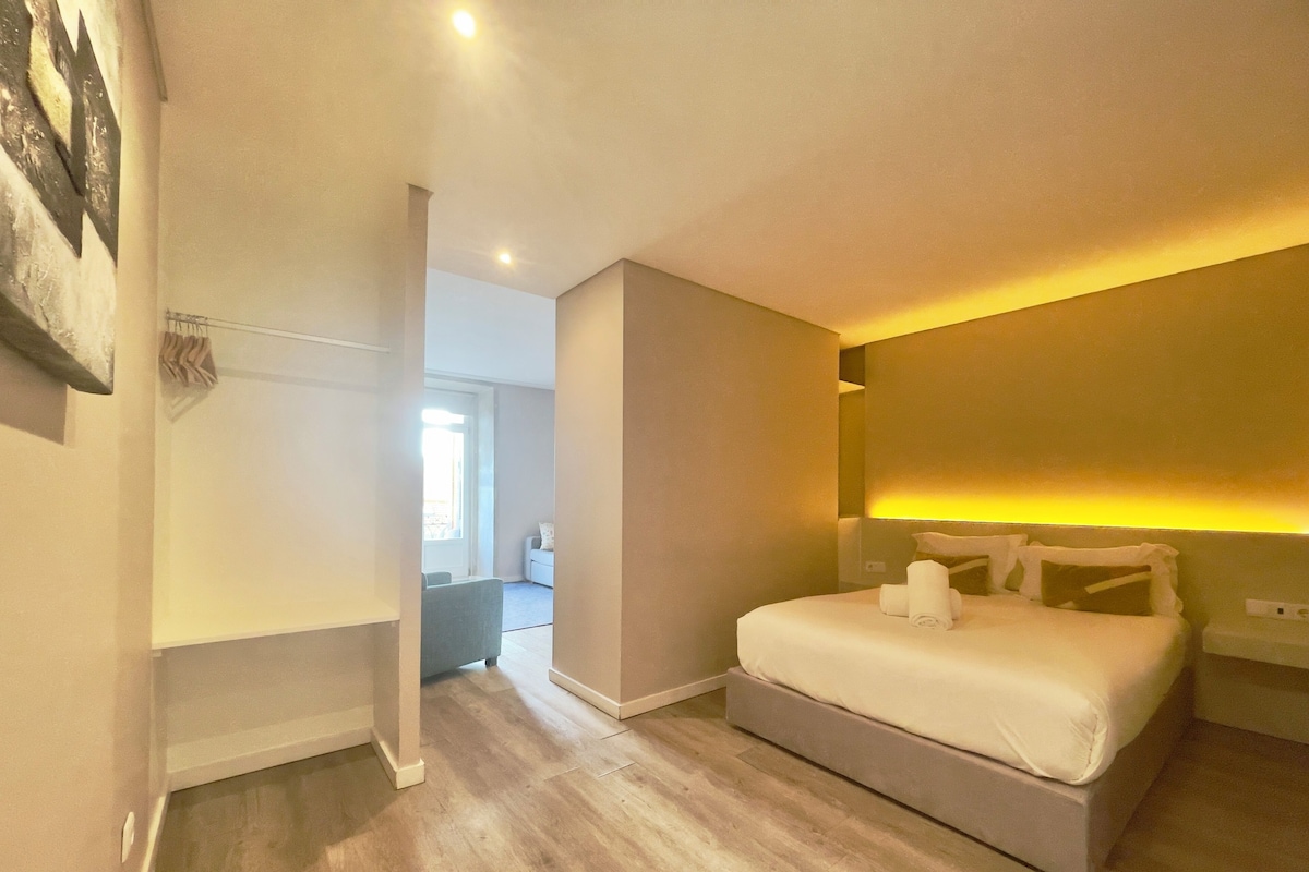 Ein modernes Hotelzimmer mit einem großen Bett, einem Nachttisch und einem kleinen Tisch daneben. Im Hintergrund ist ein Badezimmer zu sehen.