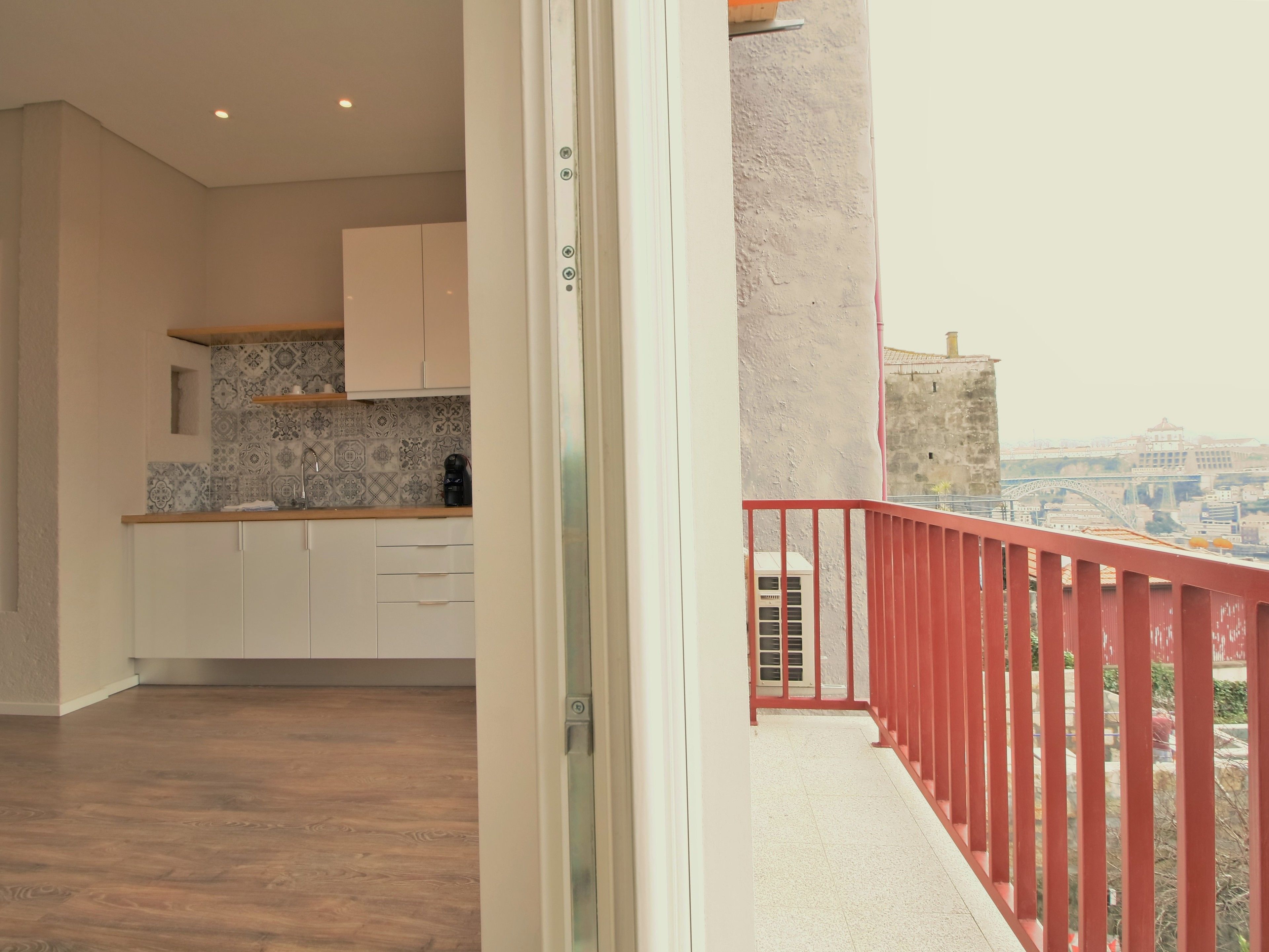 A modern kitchen with white cabinets and a patterned backsplash, leading to a balcony with a red railing overlooking a cityscape.
