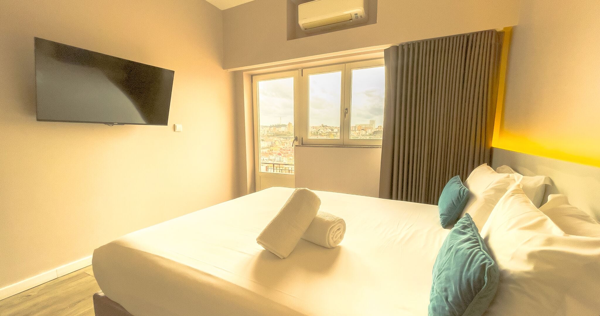 A hotel room with a bed, a TV mounted on the wall, a window with a view of the city, and a curtain.