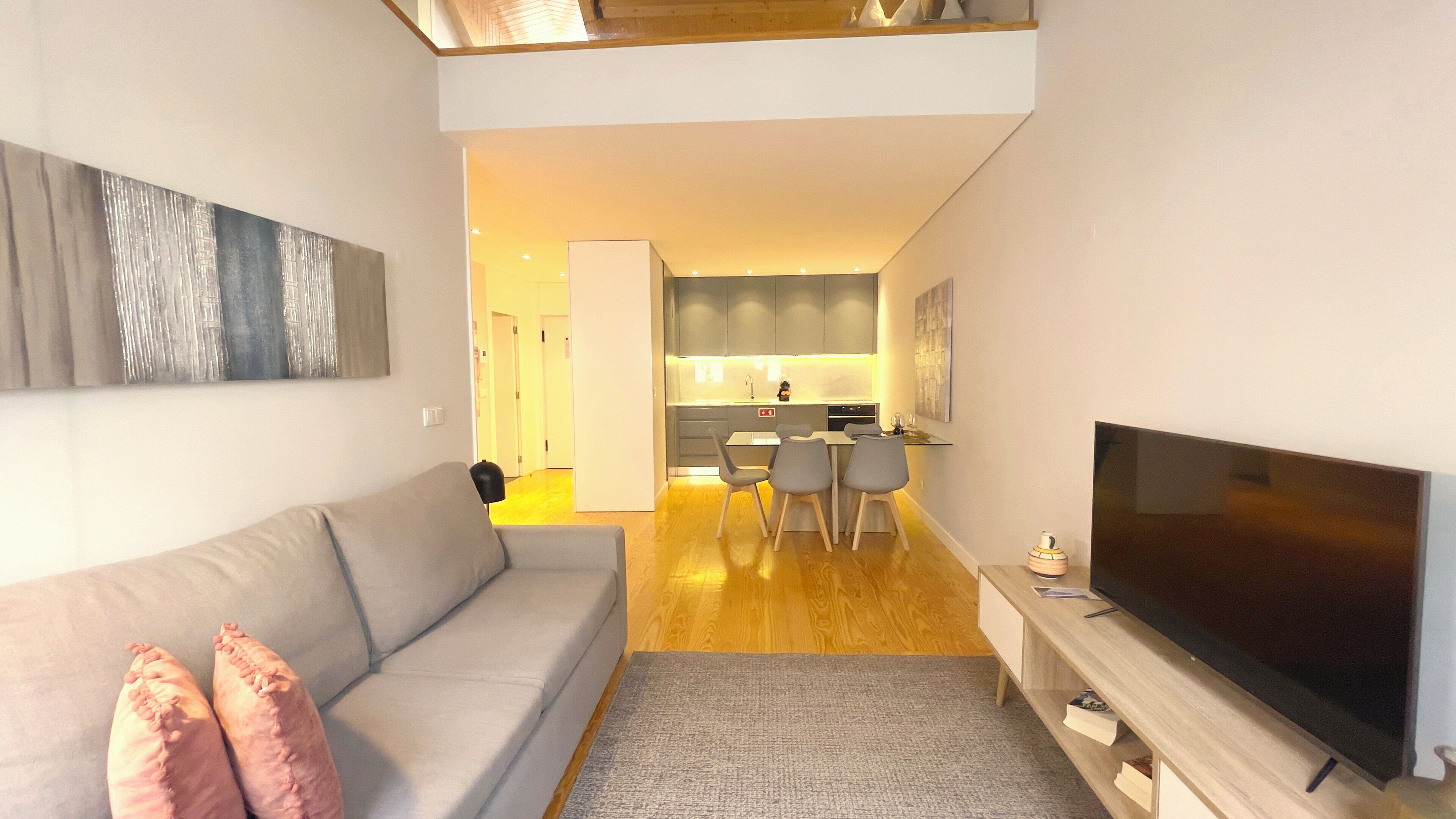 Una sala moderna con un sofá gris, un televisor de pantalla plana y un comedor con sillas, con una cocina al fondo.