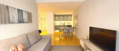 Una sala moderna con un sofá gris, un televisor de pantalla plana y un comedor con sillas, con una cocina al fondo.