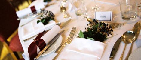 Una mesa dispuesta para un evento formal, con mantel blanco, cubertería dorada y pequeños arreglos florales.
