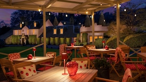 An outdoor patio with wooden tables and chairs, string lights, and a view of a house with a conical roof.