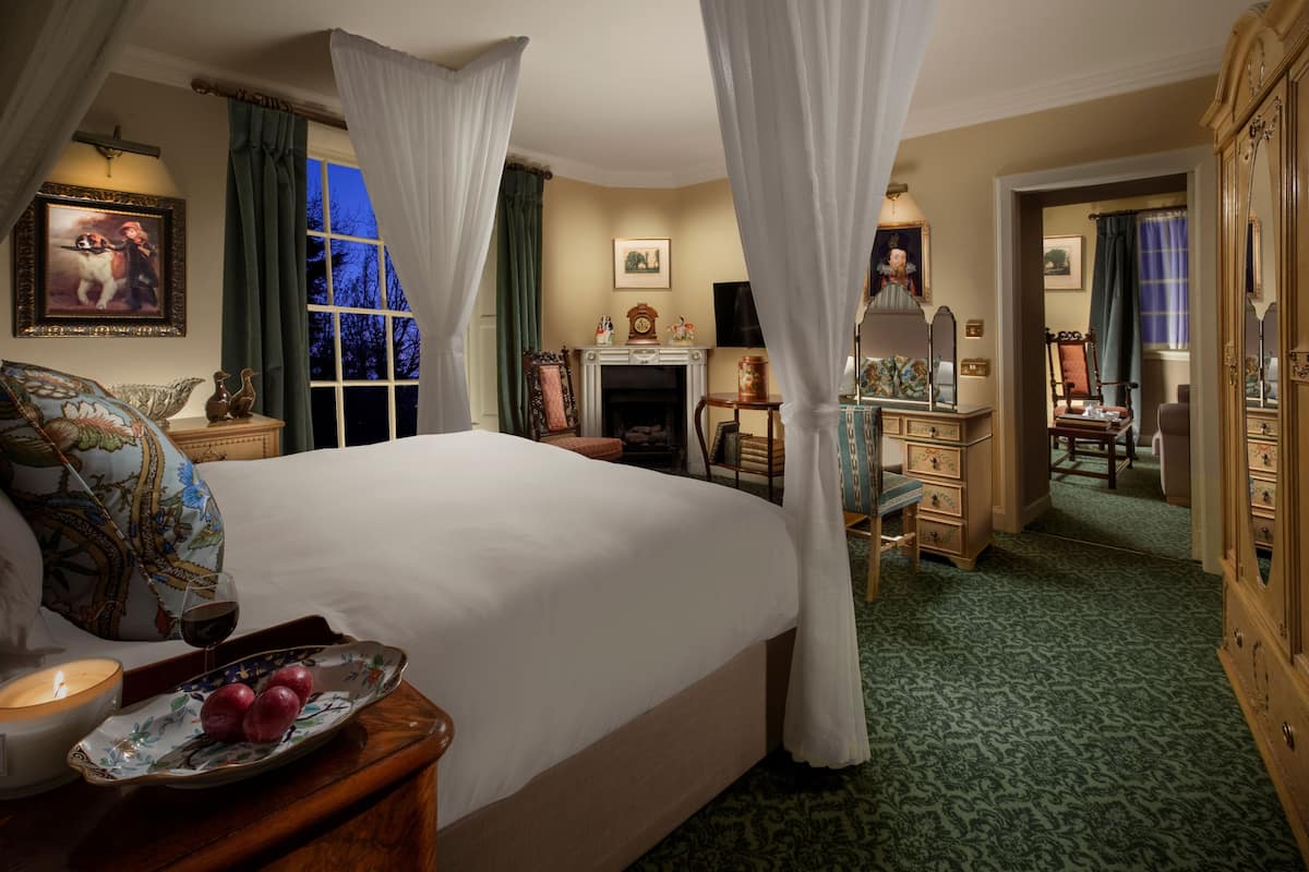 A bedroom with a large bed, a bedside table with a candle and fruit, a fireplace, and a mirror.