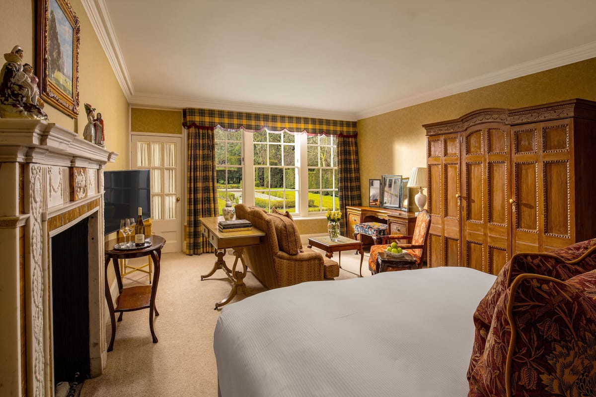 A hotel room with a bed, a desk, a chair, a wardrobe, a fireplace, and a window with a view of greenery.