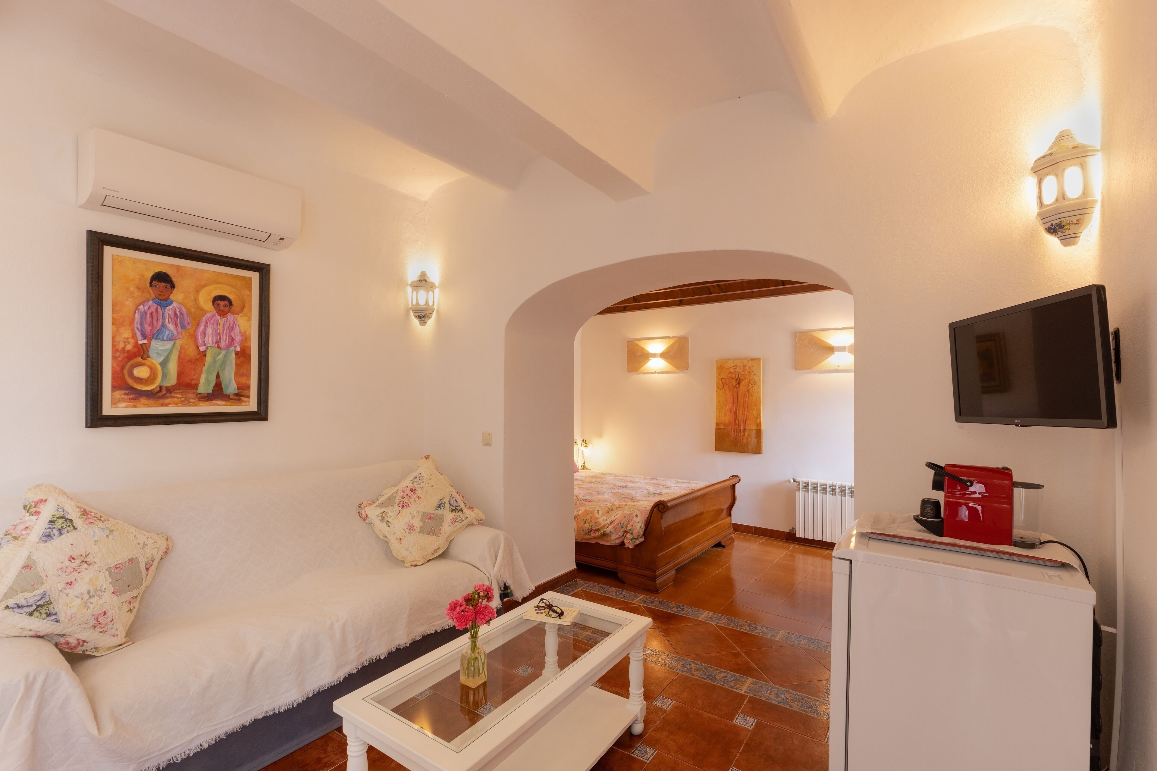 Quarto aconchegante com sofá branco, uma mesinha com um vaso, cama, televisão e um eletrodoméstico vermelho.