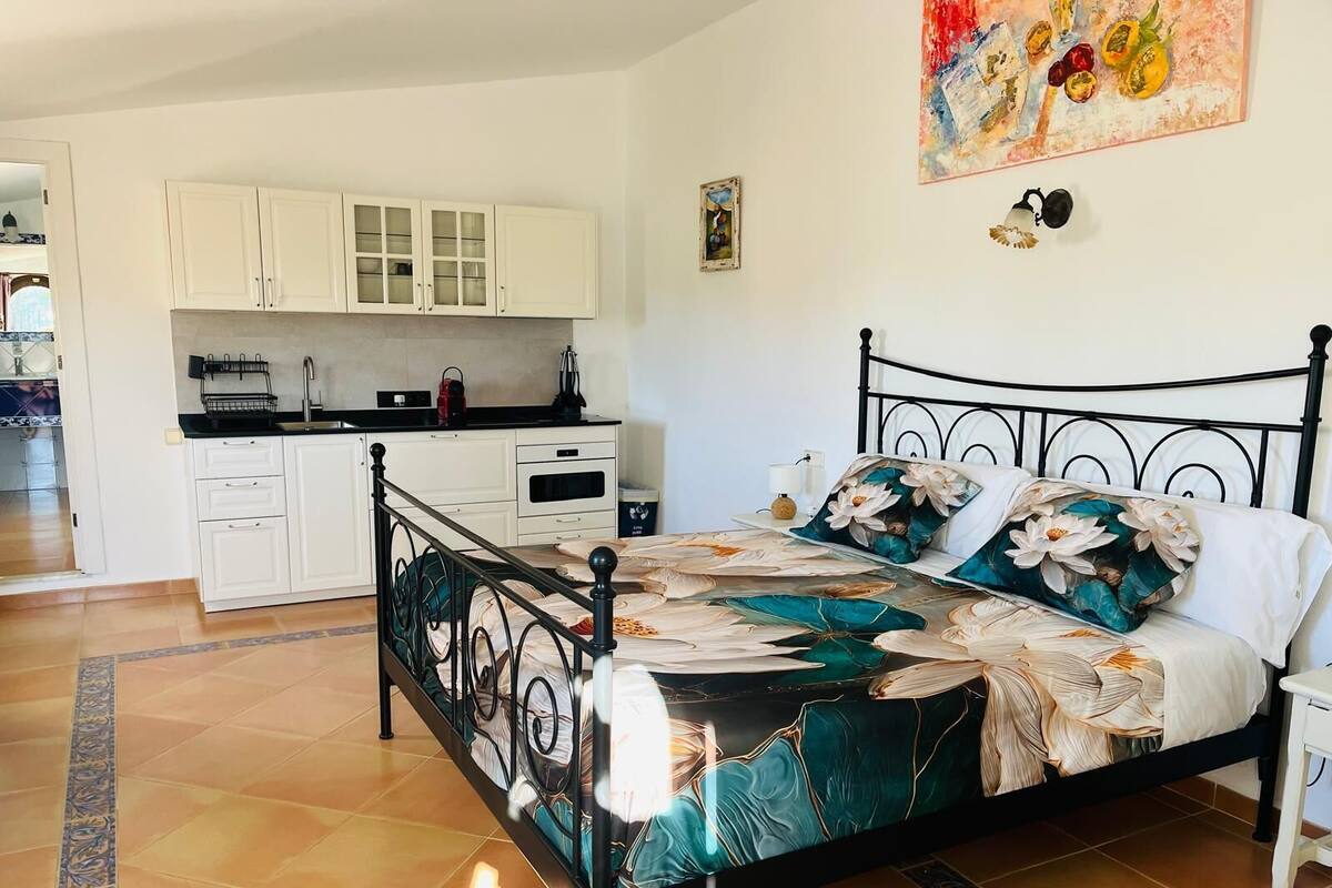 A bedroom with a bed, a nightstand, and a view of the kitchen.