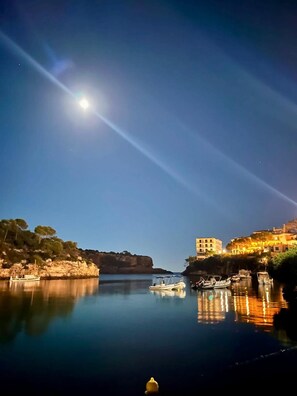 A calm waterfront at night with boats docked, a clear sky, and buildings on the shore.