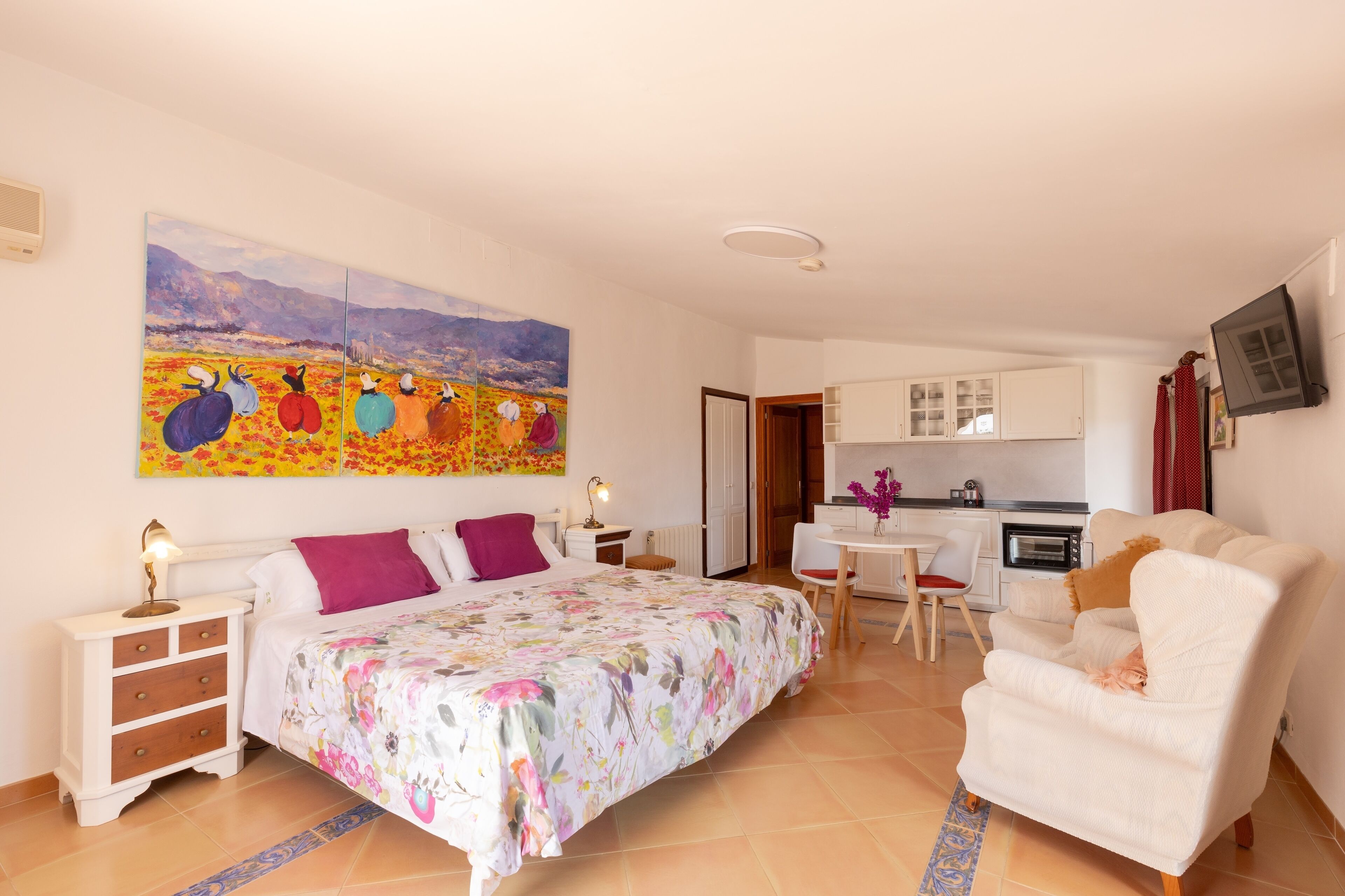 Un dormitorio con cama, mesita de noche, silla, sofá, un cuadro, una zona de cocina y un televisor.