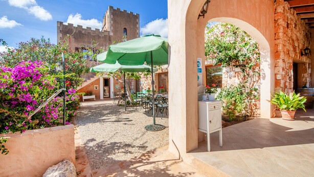 An outdoor patio area with tables and chairs, a stone wall, and a view of a castle-like building in the background. - Finca Son Marimon - Adults Only (Santanyi)