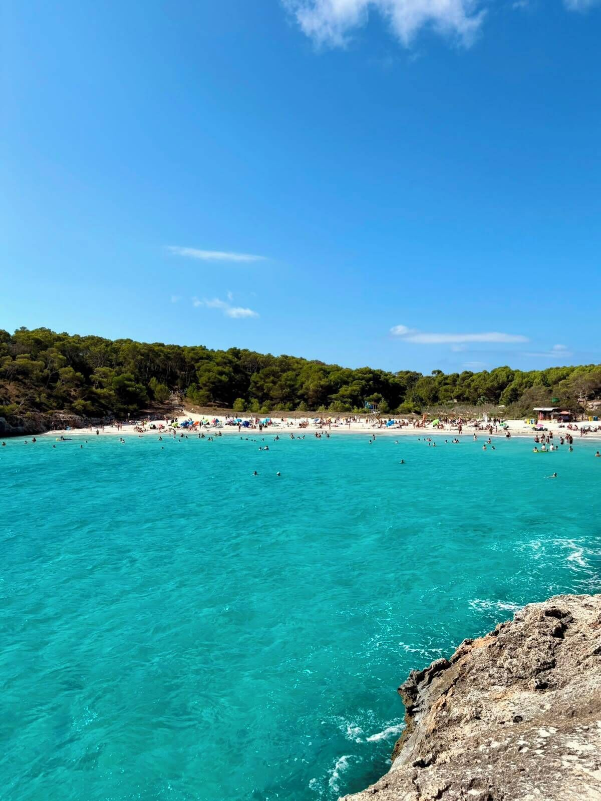 Una playa con agua turquesa cristalina, una costa rocosa y una colina boscosa al fondo.