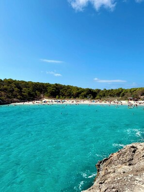 A beach with clear turquoise water, a rocky shoreline, and a forested hill in the background.