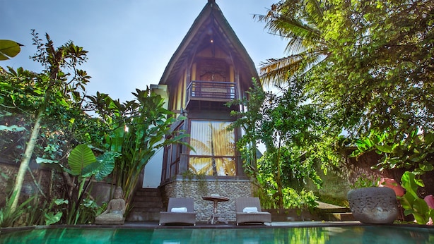 A traditional wooden building with a thatched roof, surrounded by lush greenery and a clear pool area with lounge chairs. - Gusde Tranquil Villas by EPS (Ubud)