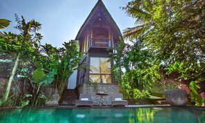 A traditional wooden building with a thatched roof, surrounded by lush greenery and a clear pool area with lounge chairs. - Gusde Tranquil Villas by EPS (Ubud)