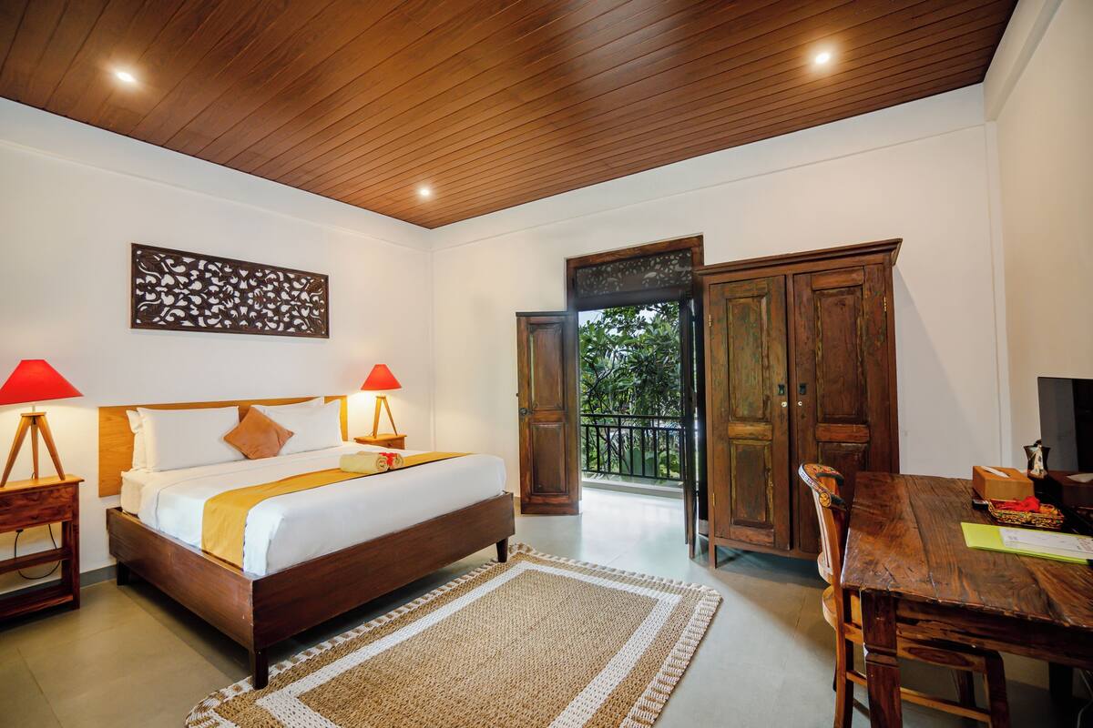 A bedroom with a wooden bed, two bedside lamps, a large window, and a wooden dining table with chairs.