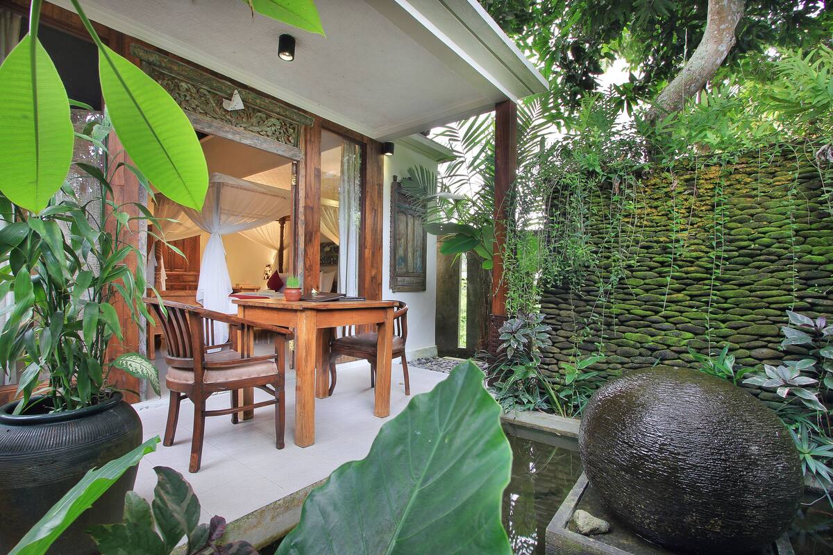 A wooden dining table with chairs, surrounded by large green plants and a water feature.