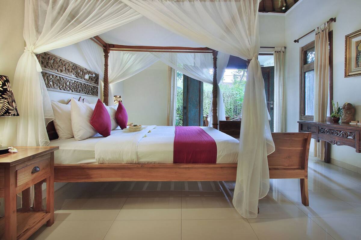 A four-poster bed with a canopy, a wooden nightstand, a desk, and a view of greenery outside.