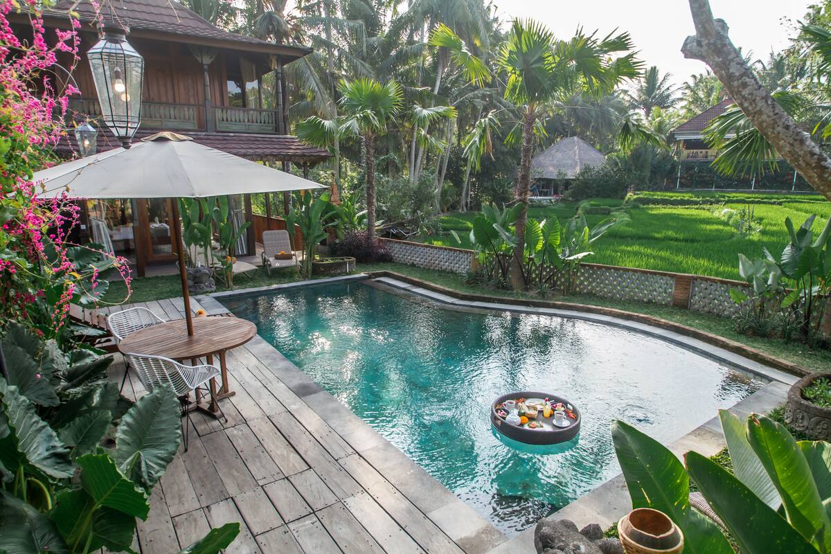 A wooden deck with a pool, surrounded by lush greenery and a thatched-roof structure.